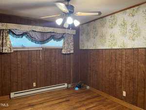 A small room with wood-paneled walls, one wall partially covered in floral wallpaper, hardwood flooring, a ceiling fan with lights, and a window with ruffled curtains. Some cables and a power strip are on the floor.