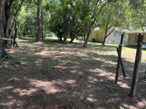 A mostly bare backyard with patchy grass, some tree cover providing shade, and a partial wooden fence in disrepair. In the background, there are two single-story brick houses and an expanse of green lawn.