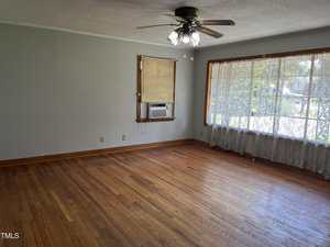 Empty room with hardwood floors, light gray walls, a ceiling fan with lights, a window with an air conditioning unit, and a large window with sheer curtains letting in natural light.