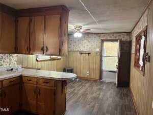 A kitchen with wooden cabinets, marble-pattern countertops, and wood-paneled walls leads into a dining area with a ceiling fan, door to the outside, and floral wallpaper. The floor is dark wood laminate.