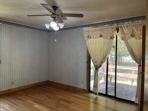Empty room with hardwood floors, striped wallpaper, and a ceiling fan with lights. Large sliding glass doors covered with sheer curtains open to a wooden deck outside. Natural light fills the space.