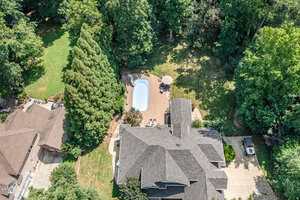 Aerial view of a house with a shingle roof, a backyard pool surrounded by a brick patio, lounge chairs, a table with an umbrella, tall trees, and a driveway with parked vehicles.