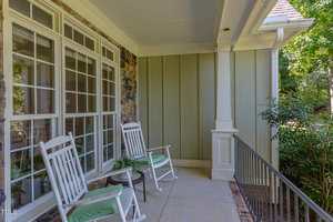 A shaded front porch with two white rocking chairs, a small table with a potted plant, large windows, stone and paneled siding, and a metal railing overlooking green plants.