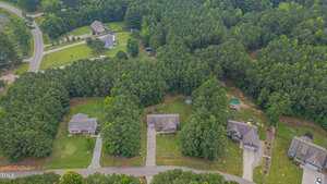 Aerial view of a suburban neighborhood with three houses along a street, large front yards, driveways, and dense trees surrounding the properties; additional houses and roads are visible in the background.