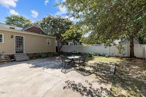 A concrete patio with a round glass table and four metal chairs is in a fenced backyard with trees and shrubs, next to a beige house with steps leading to a side door.
