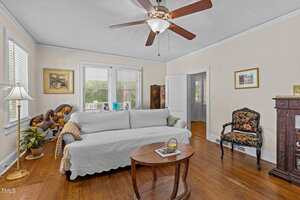 A living room with a white sofa covered by a slipcover, a patterned armchair, a wooden coffee table with books, a ceiling fan, a fireplace mantel, potted plants, framed art, and hardwood floors.