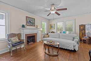 A living room features a white sofa with a white cover, a wooden coffee table, a decorative armchair, a fireplace with artwork above it, wood floors, and several windows letting in natural light.