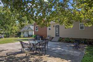 A backyard with a concrete patio featuring a round table and four chairs, additional seating in the corner, some plants along the house, and a small set of stairs leading to a door. Trees provide partial shade overhead.