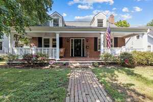 A brick house at 412 Coggeshall Street in Oxford features a large covered front porch with white railings and an American flag. A brick walkway winds through landscaped bushes and flowers, leading to the inviting front steps under a blue, cloud-streaked sky.