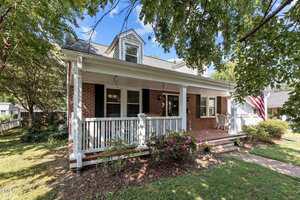 A two-story brick house with a covered front porch, white columns, and railing. There are bushes and flowering plants in the front yard, and an American flag hangs near the porch. Trees provide shade around the house.