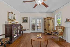 A living room with wood floors, a ceiling fan, a round coffee table, an ornate cabinet, two upholstered chairs, a large armoire, French doors, and framed art on cream-colored walls.