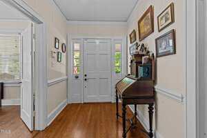 A hallway with wooden floors, beige walls, white trim, and crown molding. A white front door with sidelights lets in natural light. A wooden writing desk and framed pictures are on the right wall.