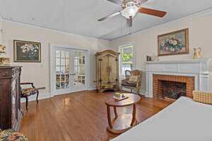 A living room with wood floors, a ceiling fan, a fireplace with a brick interior, an ornate armoire, a wooden coffee table, a cushioned bench, and floral artwork on the walls. French doors lead to another room.