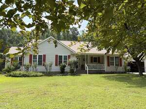 A single-story house with beige siding, red shutters, and a covered front porch sits on a well-maintained lawn with trees and shrubs in the yard.