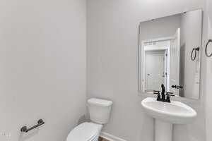 A small bathroom with a white toilet, a white pedestal sink, a rectangular mirror above the sink, a wall-mounted towel ring, and a toilet paper holder. The walls are light gray.