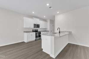 Modern kitchen with white cabinets, stainless steel appliances, gray granite countertops, a center island with a black faucet, and light wood flooring. The walls are painted white and the room is brightly lit with ceiling lights.