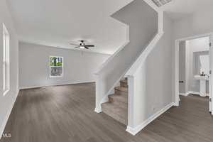 A modern, unfurnished living space with light gray walls, wood flooring, a carpeted staircase, a ceiling fan, and natural light from two windows; a bathroom is visible through an open door on the right.