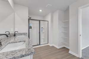 A modern bathroom with a double sink vanity, granite countertop, a glass-enclosed shower, built-in wall shelves, and wood-look flooring. An open doorway leads to a carpeted room.