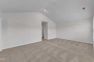 Empty room with light beige carpet, white walls, a vaulted ceiling, and a doorway leading to another area; overhead lighting and a ceiling vent are visible.