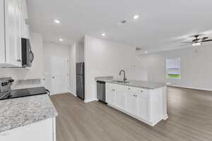 A modern kitchen with white cabinets, stainless steel appliances, granite countertops, and light wood flooring, open to a living area with a ceiling fan and a window. The space is unfurnished and well-lit.