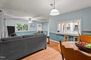 A living room with light blue walls, wood flooring, a gray sectional sofa, wall-mounted TV, ceiling fan, and large windows; a dining table and a breakfast bar with stools are also visible.