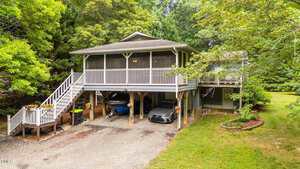A two-story house with a screened porch and white railings above a carport, which shelters two vehicles and storage items. The home is surrounded by trees and greenery, with a gravel driveway in front.