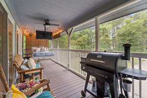 A covered porch with ceiling fan, string lights, a hanging daybed, two wooden chairs with cushions and potted flowers, and a black grill, overlooking a wooded yard.