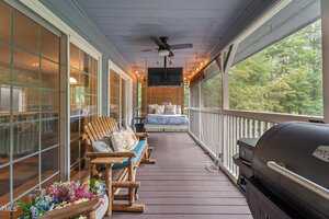 A covered screened porch with wooden flooring, a ceiling fan, string lights, a porch swing bed with pillows, a wooden bench and chairs, potted flowers, a grill, and large windows overlooking trees.