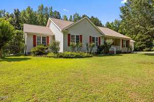 Single-story house with light-colored siding, red shutters, and a covered front porch, surrounded by green grass and trees under a clear blue sky. Shrubs and small plants line the front of the home.