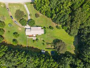 Aerial view of a house with a light-colored roof surrounded by green grass, trees, a driveway with parked cars, a backyard garden area, and a wooded area bordering the property.