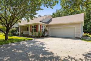 Single-story house with beige siding, a front porch with white railings, red shutters, a double garage, and a concrete driveway. Trees provide shade in the front yard, and the sky is clear and blue.