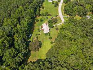 Aerial view of a house surrounded by large grassy lawns and dense trees, with a curved road and a few cars nearby; other houses and open spaces are visible in the surrounding area.