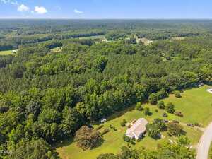 Aerial view of a house with a large yard surrounded by trees and forested areas, with a few neighboring homes visible and expansive greenery extending into the distance under a blue sky.