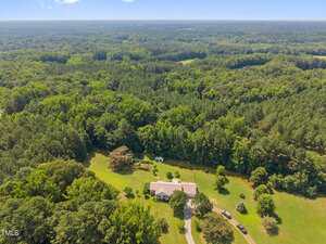 A large house with a white roof sits on a spacious, grassy lot surrounded by dense forest. A long driveway leads to the house, and several outbuildings and trees dot the property. Rolling hills and trees fill the background.