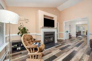 A living room with a rocking chair, fireplace with a TV above it, a potted plant, a floor lamp, and wood flooring. An open doorway leads to a dining area in the background.