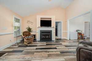 Living room with light peach walls, a fireplace under a wall-mounted TV, a wicker chair with a cushion, large windows, and multi-toned wooden flooring. Doorways lead to adjacent rooms.