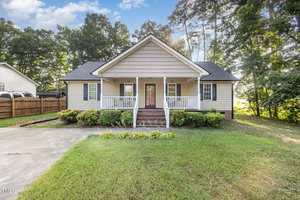 Single-story house with beige siding, a covered front porch, white railings, and steps leading to the entry door. Shrubs line the front, with a lawn and trees surrounding the property. The driveway is visible on the left.