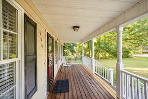 A covered front porch with wooden flooring, white railing, beige siding, black door with house number 609, a small chair, a black doormat, and trees and grass visible in the yard.