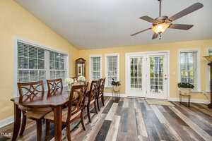 A dining room with a long wooden table and eight chairs, wood-look flooring, yellow walls, ceiling fan with light, multiple windows, and double glass doors leading outside. A potted plant and mirror are in the background.