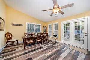 A dining room with a wooden table and six chairs, wood-look tile flooring, yellow walls, three large windows, a glass display cabinet, a ceiling fan with a light, and double glass doors leading outside.