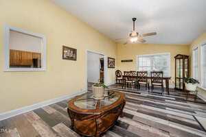 A living and dining area with yellow walls, patterned wood flooring, a glass-top coffee table, a dining table with chairs, wall art, and large windows letting in natural light. A ceiling fan is on the ceiling.