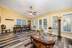 A bright dining room with yellow walls, a ceiling fan, and large windows. A wooden dining table with chairs sits near the windows, and a coffee table with a potted plant is centered on a patterned floor.