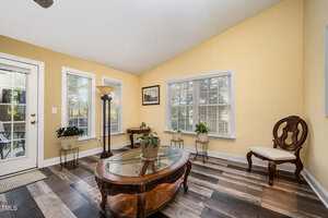A small sunroom with yellow walls, large windows, and wood-patterned flooring. The room contains a glass coffee table, potted plants, a standing lamp, and two wooden chairs with white cushions.