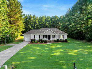 Single-story white house with a front porch, attached garage, and neatly trimmed shrubs, set on a large green lawn surrounded by tall pine trees under a blue sky.