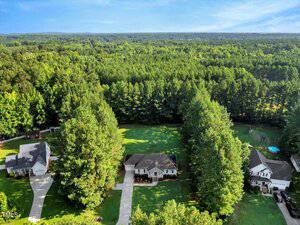 Aerial view of three houses with large yards, surrounded by dense green trees and forest under a clear blue sky, with long driveways leading to each house.