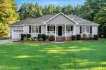 Single-story house with light-colored siding, black shutters, and a covered front porch. There is a two-car garage on the left, trimmed bushes along the front, and a large, well-maintained lawn with trees in the background.