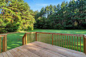 A wooden deck with railings overlooks a green lawn bordered by dense, tall trees under a clear blue sky.
