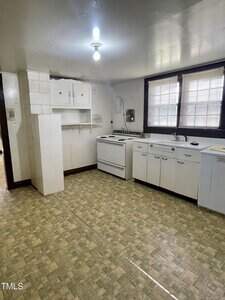 A small kitchen with white cabinets, a white stove, a sink under a window with blinds, and a section of open shelving. The floor has a patterned vinyl covering and the ceiling has a single exposed light bulb.
