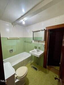 Small bathroom with light green tile walls, a white bathtub, a white sink with a mirror above it, a white toilet with a beige seat, and a patterned green floor. A single exposed bulb lights the room.