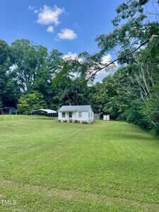 A small white house with a green roof sits in the center of a large grassy yard, surrounded by trees. A metal carport and a small shed are visible in the background under a blue sky with scattered clouds.